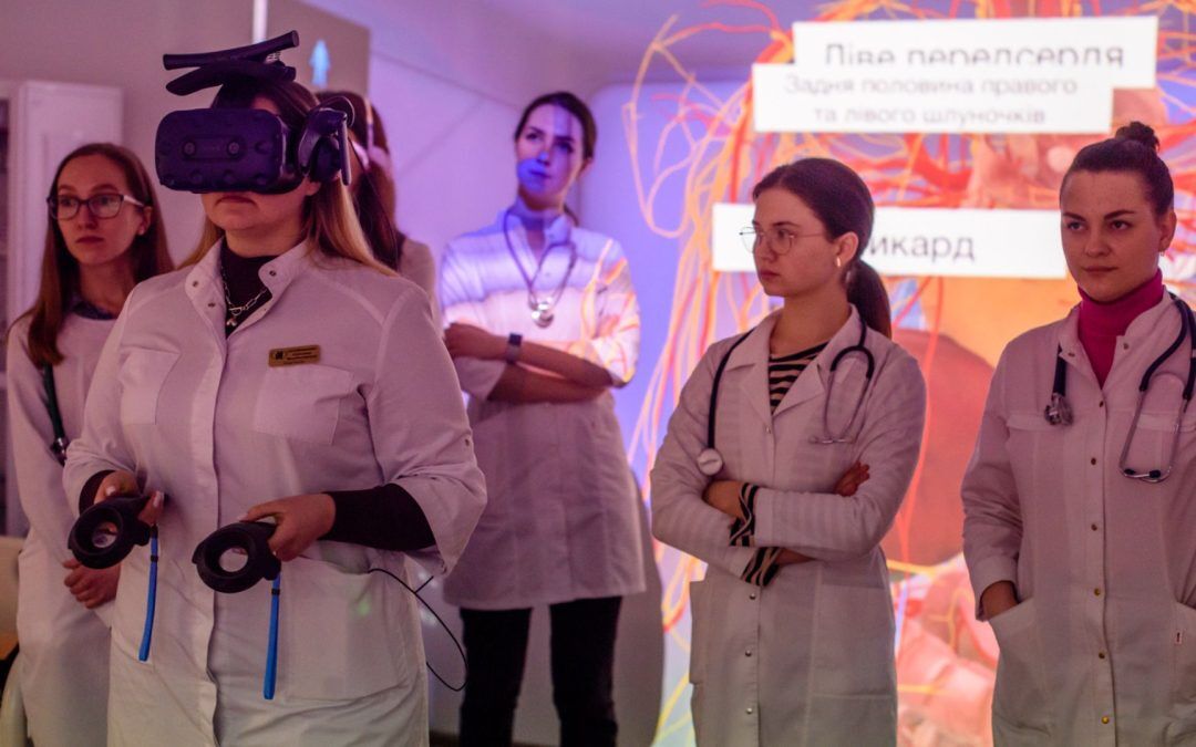 A group of medical students at Zaporizhzhia State Medical and Pharmaceutical University (ZSMPhU) participate in an immersive anatomy lesson. One student wears a VR headset and holds motion controllers, engaging with 3D anatomical content, while others observe attentively. The background features a projected anatomical diagram in Ukrainian, enhancing the interactive learning environment. All students are dressed in white lab coats and appear focused and engaged.